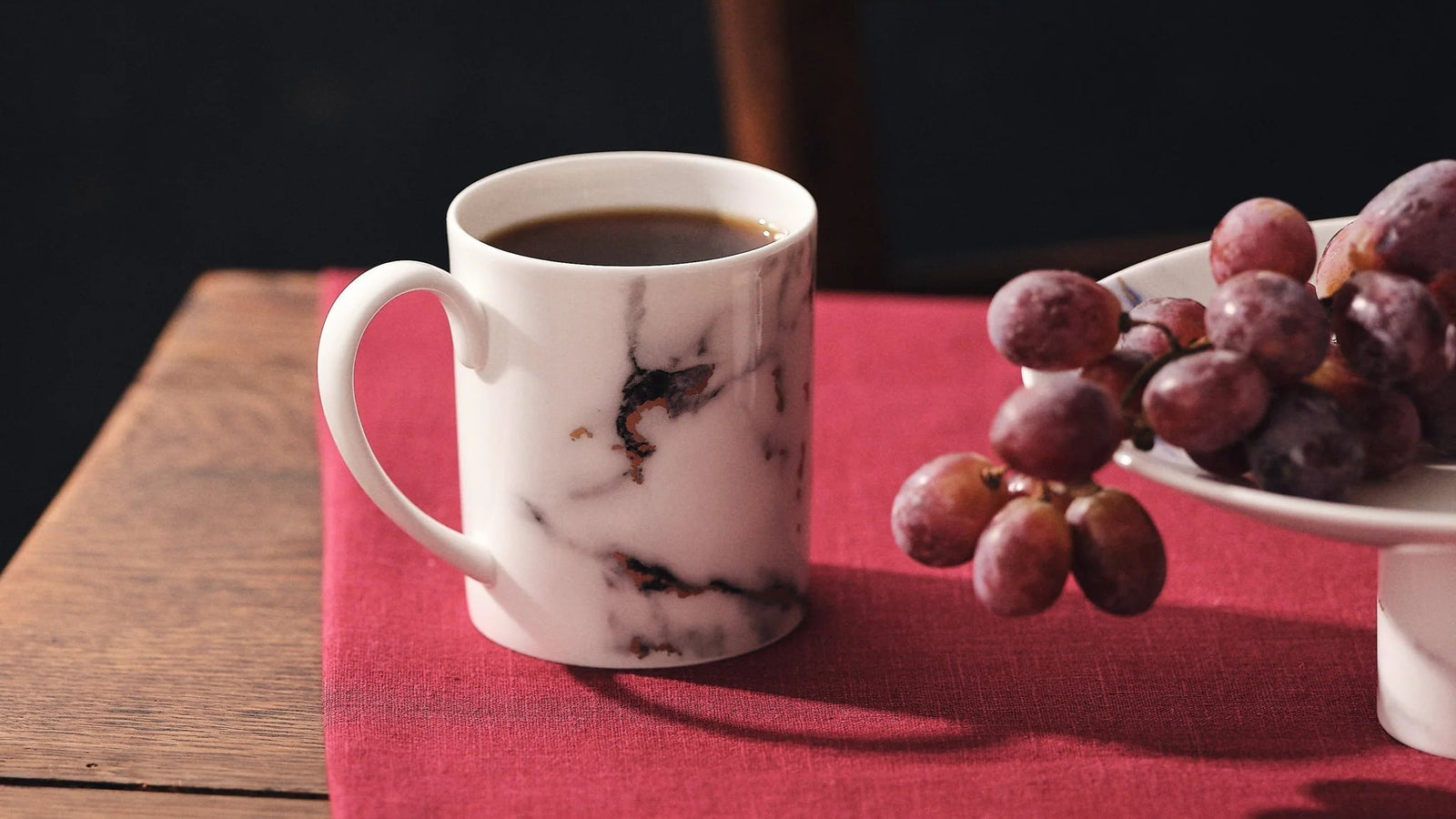 Marble-patterned mug with coffee on a red tablecloth, accompanied by grapes and a plate.